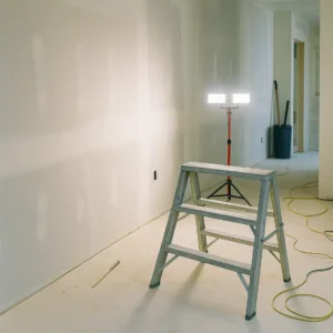 Interior view of a home under renovation with drywall installation in progress, work light and step ladder set up for wall finishing tasks.